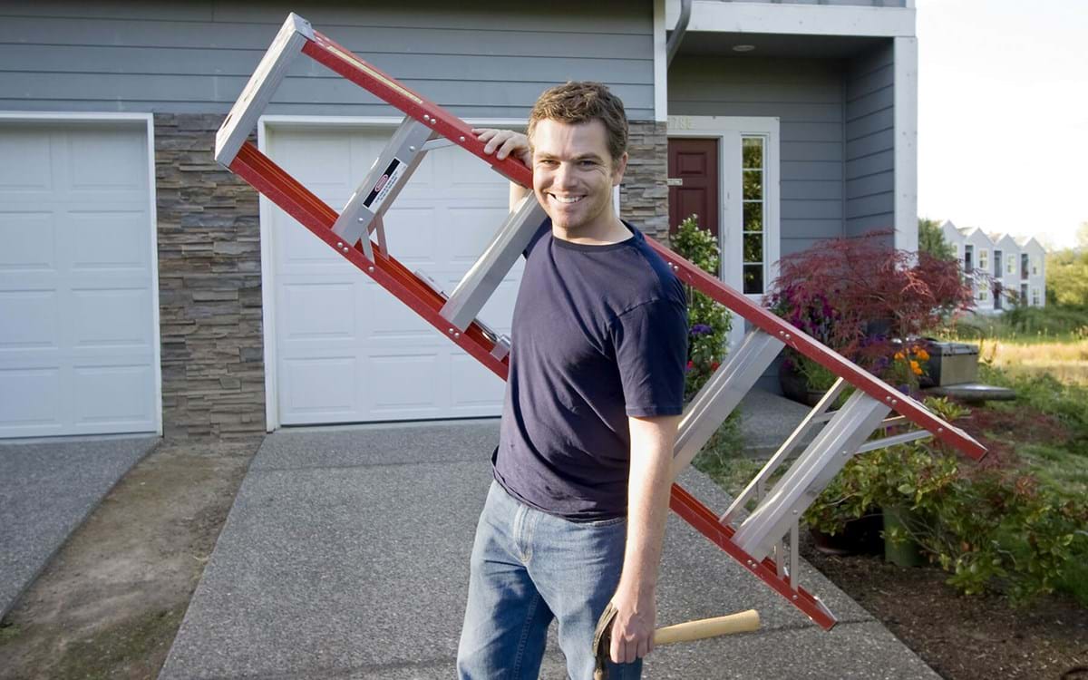Man standing in front of his house holding a ladder and a hammer